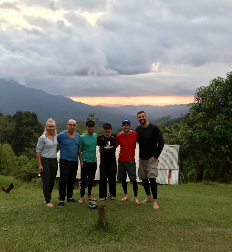 Six people in athletic wear stand together in a grassy field in front of a corrugated metal building. A rugby ball sits on the ground in front of them, and a dramatic sunset glows behind a deep mountain range in the background.