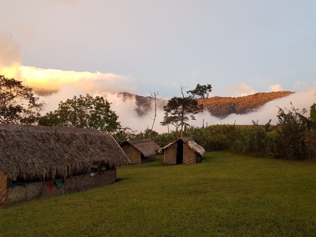 Simple thatched-roof huts sit in a green grassy clearing as thick white clouds roll over a forested mountain ridge at sunset.