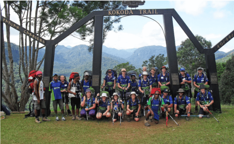 Large, diverse groups of trekkers and local guides pose at major landmarks, such as the Kokoda Trail archway, or navigate misty, high-altitude meadows.