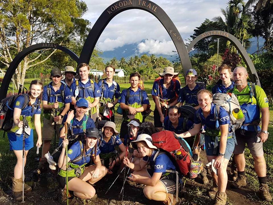 A group of young trekkers in blue and green foundation uniforms poses together at the entrance arches of the Kokoda Trail.