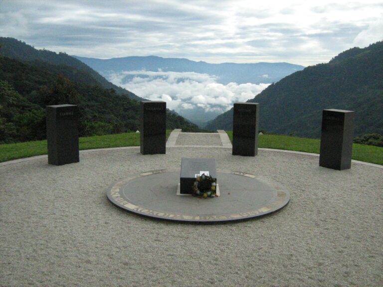 Circular memorial site in a mountainous area with lush greenery and distant hills under a partly cloudy sky; central circular platform with square pedestal surrounded by embedded ring; five upright black stone plaques arranged around the platform; two flagpoles stand on either side; bordered by grass and stone pathways, creating a solemn commemorative atmosphere.