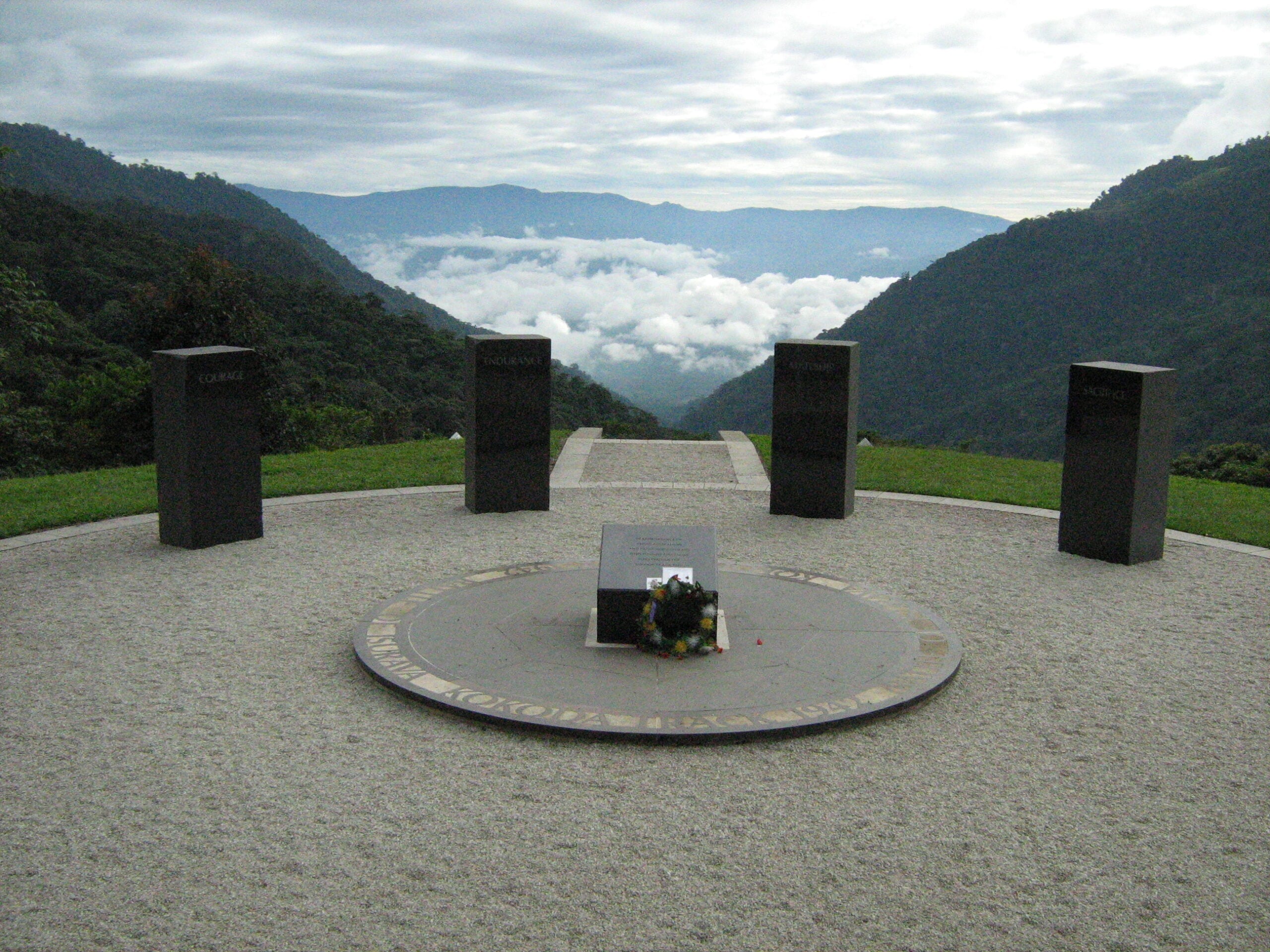 Circular memorial site in a mountainous area with lush greenery and distant hills under a partly cloudy sky; central circular platform with square pedestal surrounded by embedded ring; five upright black stone plaques arranged around the platform; two flagpoles stand on either side; bordered by grass and stone pathways, creating a solemn commemorative atmosphere.