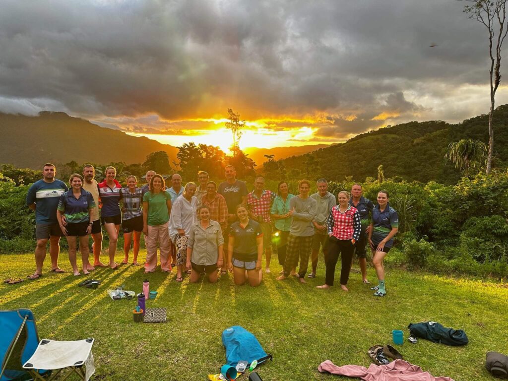 Group of twenty people standing and kneeling on grassy area with outdoor gear scattered nearby; lush green hills in background; sunset casts golden light through clouds and trees, creating dramatic and picturesque atmosphere; scene conveys camaraderie and adventure in natural setting.