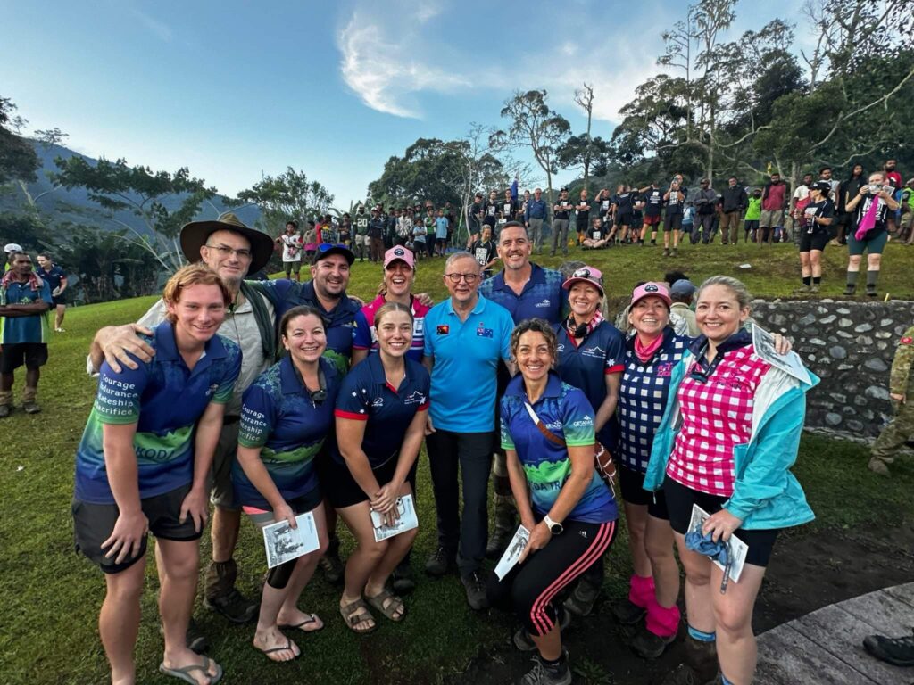 Group of thirteen people posing outdoors in a lush mountainous area; many wear matching shirts with words “Courage,” “Endurance,” “Mateship,” and “Sacrifice” printed on sleeves, suggesting commemorative or team event; some hold booklets; larger crowd with numbered bibs gathered behind, indicating organized trek or race; setting conveys camaraderie and cultural or historical significance