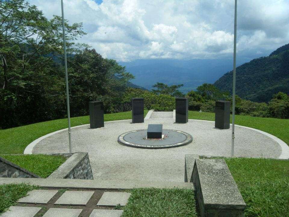 Circular memorial site in a mountainous area with lush greenery and distant hills under a partly cloudy sky; central circular platform with square pedestal surrounded by embedded ring; five upright black stone plaques arranged around the platform; two flagpoles stand on either side; bordered by grass and stone pathways, creating a solemn commemorative atmosphere.