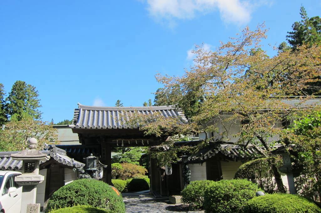 Traditional Japanese entrance gate with tiled roof and curved eaves, surrounded by manicured bushes, trees, and stone lanterns; hanging signs and stone markers with Japanese characters add cultural detail, under a clear blue sky.