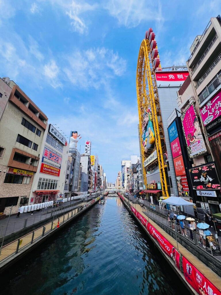 Bustling urban scene of Osaka’s Dotonbori district, viewed from a bridge over the canal; pedestrian walkways with people under umbrellas line both sides, while buildings are covered in colorful advertisements and signage, including a large yellow Ferris wheel attached to the Don Quijote store; bright atmosphere under a clear sky with scattered clouds.