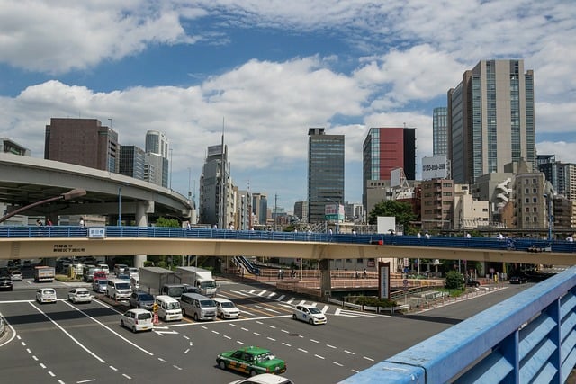 A high-angle view of a busy city intersection in Tokyo with a pedestrian bridge in the foreground, an elevated highway on the left, and various modern skyscrapers under a blue, cloudy sky.