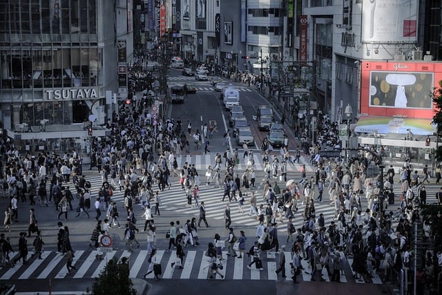 A high-angle view of a massive crowd of pedestrians crossing a multi-directional zebra-stripe crosswalk in a busy urban intersection surrounded by tall buildings and city traffic.