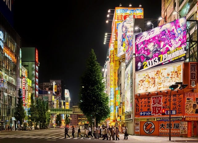 A vibrant night view of a busy street in Akihabara, Tokyo, with pedestrians crossing a crosswalk and multi-story buildings covered in glowing neon signs and large anime advertisements.