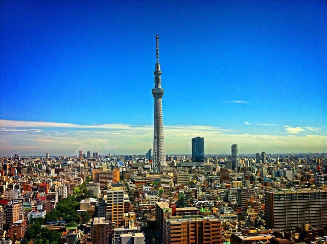 A wide aerial view of the Tokyo city skyline under a clear blue sky, dominated by the tall, silver Tokyo Skytree tower rising from the center of a dense urban landscape.