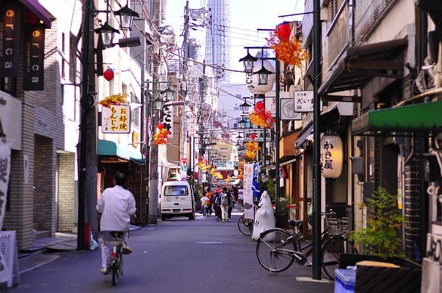 A narrow, busy city street in Japan lined with shops, banners, and lanterns, with a person riding a bicycle away from the camera and a tall tower visible in the distant background.
