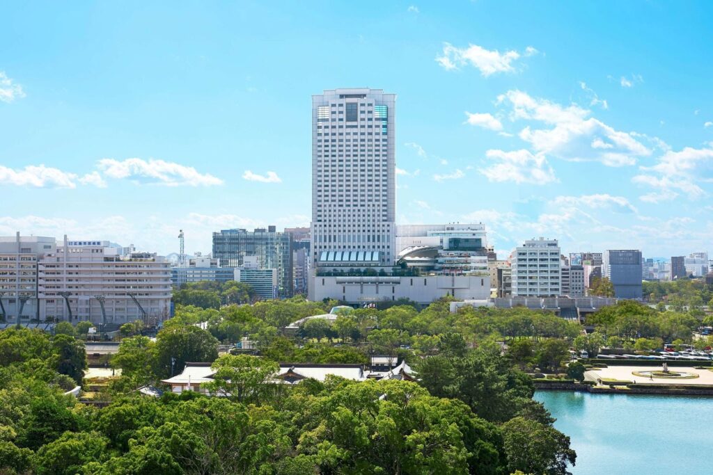 Cityscape with a tall modern high‑rise building at the center, surrounded by mid‑rise structures; foreground features a lush green park with dense trees and a body of water, under a sunny sky with scattered clouds.