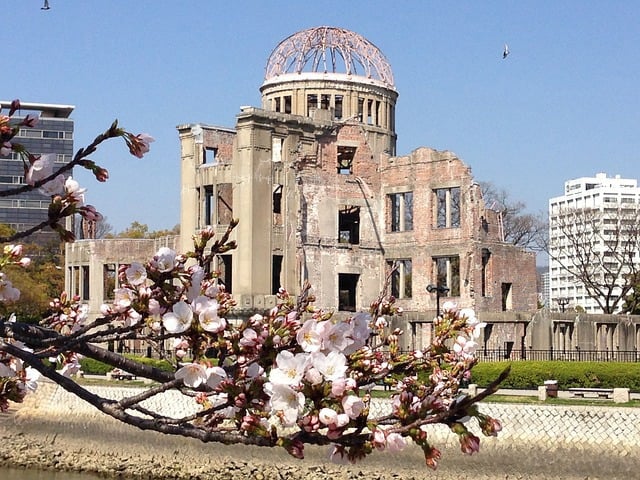 A view of the ruined Hiroshima Atomic Bomb Dome framed by blooming cherry blossoms in the foreground, with a river and city buildings under a clear blue sky.