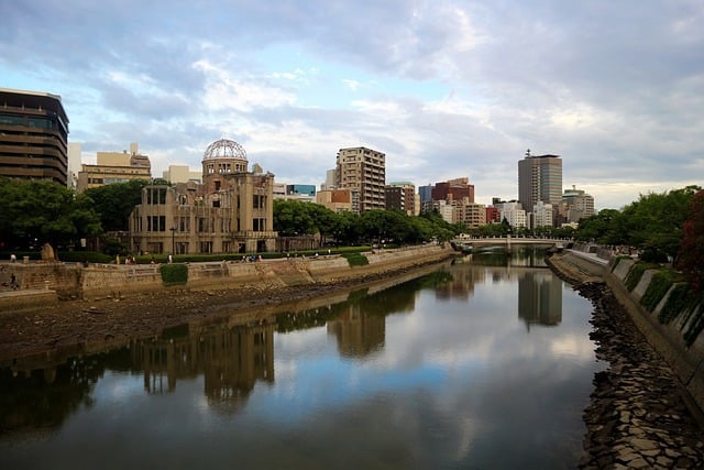 A wide-angle view of the Atomic Bomb Dome standing along a riverbank, reflected in the calm water, with a modern city skyline and a bridge in the background under a cloudy sky.