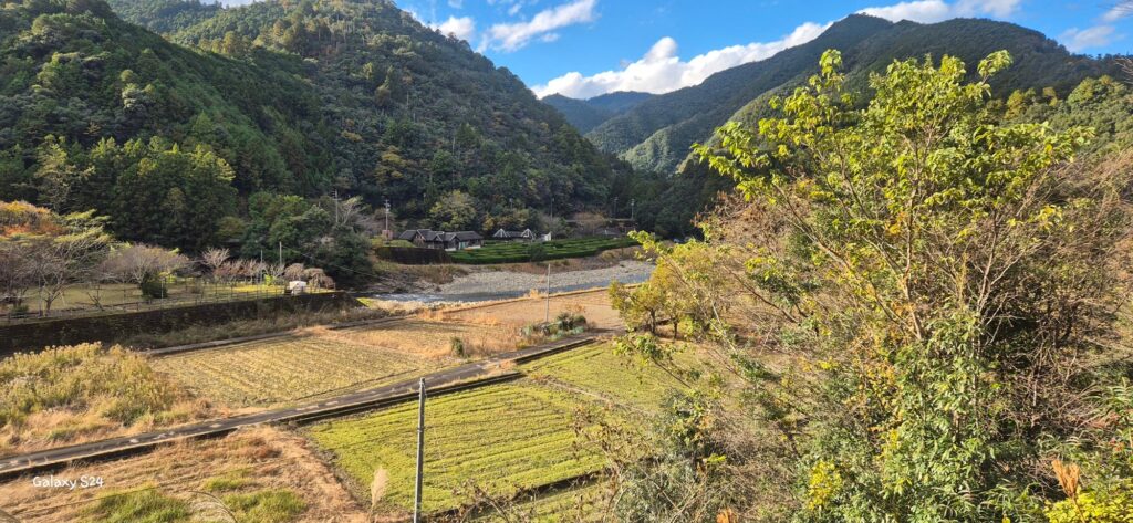 Scenic rural landscape with terraced fields in the foreground, a small village of traditional houses at the base of lush green mountains, and a riverbed running through the scene; partly cloudy sky with patches of blue adds to the tranquil atmosphere.