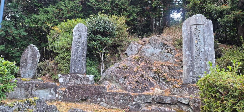 Group of vertical stone monuments with Japanese inscriptions set among rocks and greenery in a forested area; commemorative steles suggesting cultural or historical significance.