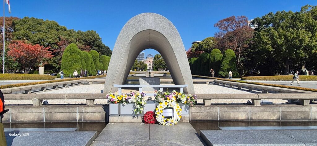 Hiroshima Peace Memorial Park with the saddle‑shaped Cenotaph for A‑bomb Victims in the foreground, adorned with floral tributes; aligned through the arch is the Atomic Bomb Dome, a preserved ruin symbolizing the devastation of the atomic bomb; landscaped grounds with trimmed trees, pathways, and visitors walking nearby.