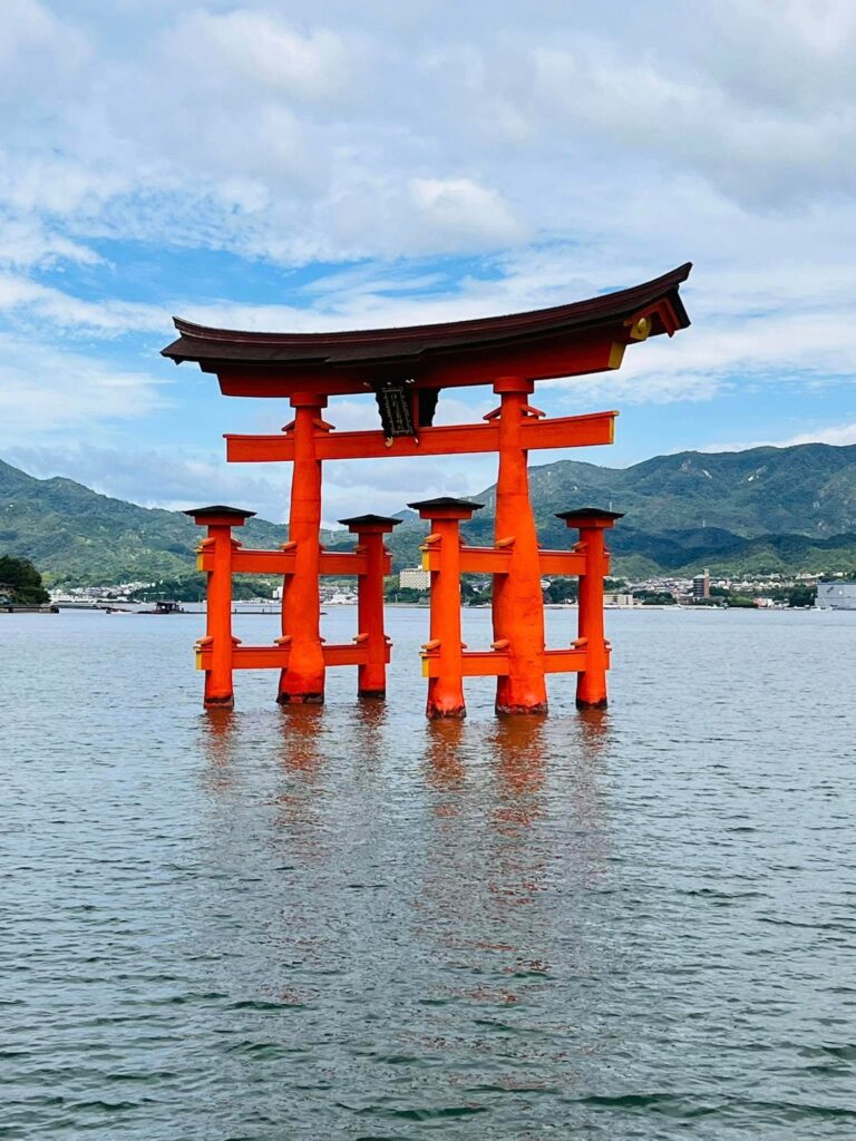 Iconic floating vermilion torii gate of Itsukushima Shrine standing in the water, with lush green mountains and a small town in the background under a partly cloudy sky.