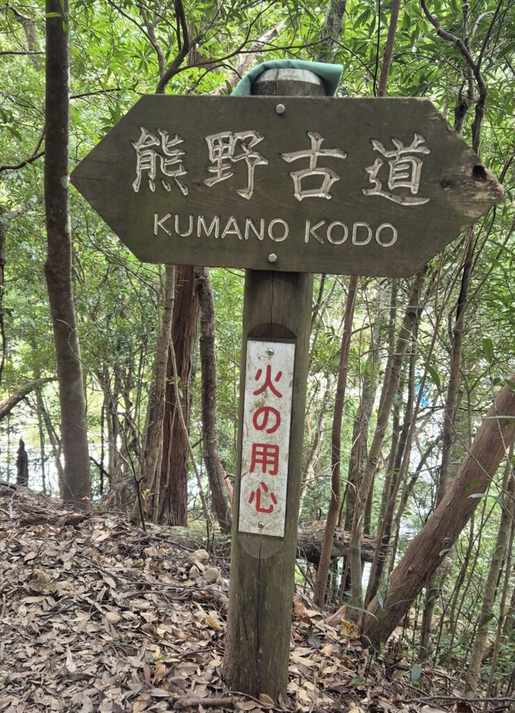 Wooden arrow‑shaped signpost in a forested area with Japanese text “熊野古道” and English translation “KUMANO KODO”; below it, a vertical sign with red Japanese text reading “火の用心” meaning “Fire caution.”