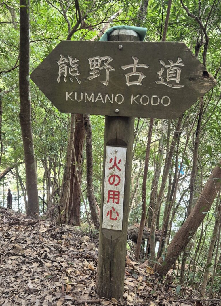 Wooden arrow‑shaped signpost in a forested area with Japanese text “熊野古道” and English translation “KUMANO KODO”; below it, a vertical sign with red Japanese text reading “火の用心” meaning “Fire caution.”