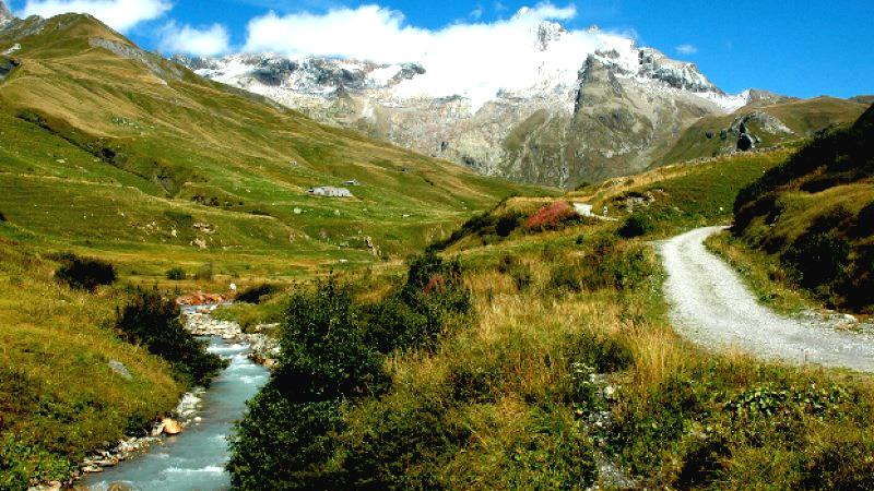 Picturesque alpine landscape with a winding gravel path beside a flowing stream, surrounded by green grass and bushes, leading toward snow-capped peaks under a partly cloudy blue sky.