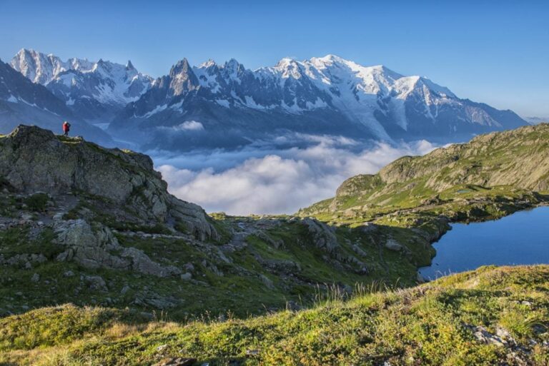A lone hiker in a red jacket stands on a rocky ridge overlooking a sea of clouds and a massive, snow-covered mountain range.