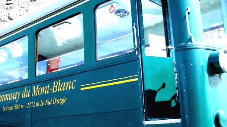 Green tram car labeled “Tramway du Mont-Blanc” with route details “Le Fayet 584 m – 2372 m Le Nid D’aigle” in yellow text, featuring large windows, open door revealing interior controls, and exterior decorated with yellow stripes and metallic handles.