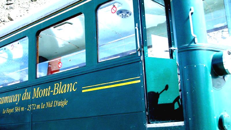 Green tram car labeled “Tramway du Mont-Blanc” with route details “Le Fayet 584 m – 2372 m Le Nid D’aigle” in yellow text, featuring large windows, open door revealing interior controls, and exterior decorated with yellow stripes and metallic handles.