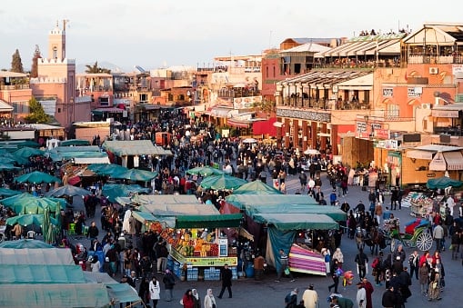A high-angle view of a crowded public square in Marrakesh, filled with rows of green-canopied market stalls and hundreds of people walking between them.