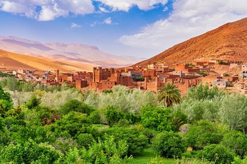 A sprawling view of a traditional earth-toned Moroccan village situated behind a dense, green valley filled with palm trees and lush vegetation, set against arid desert mountains.