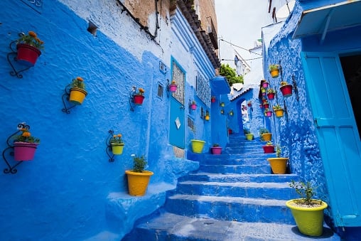A bright blue narrow alleyway in Chefchaouen, Morocco, with stone stairs leading up between high walls. Colourful flower pots in shades of yellow, pink, and red are mounted on the walls.