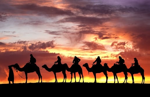 A silhouette of a line of travelers riding camels across a desert at sunset. The sky is filled with dramatic orange, purple, and yellow clouds.