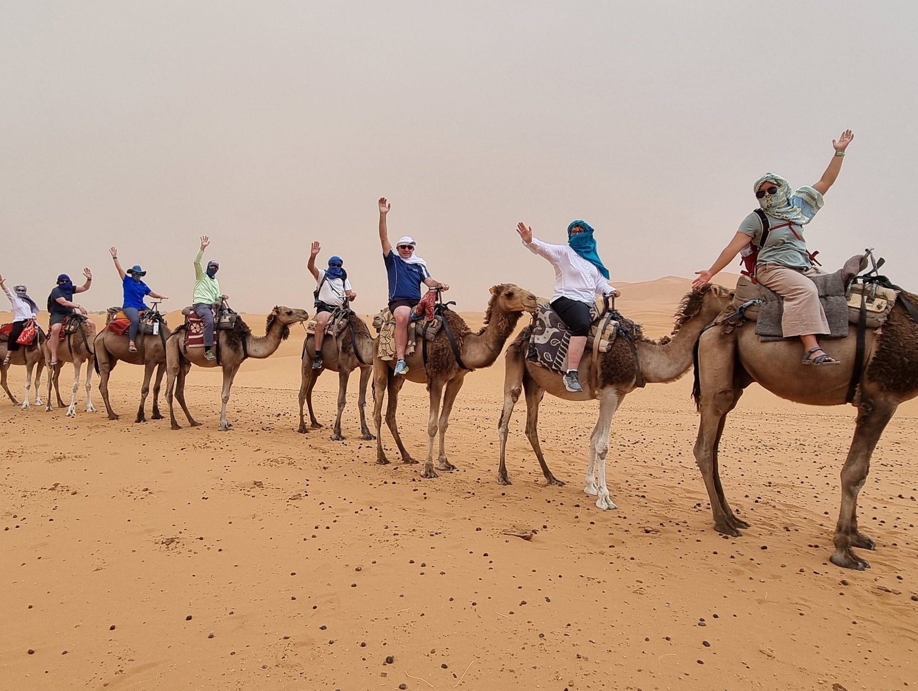 Group of seven people riding camels in desert; each rider raises one arm in gesture of excitement or celebration; camels arranged in single line; riders wear head coverings and casual clothing suited for desert; expansive sand dunes stretch into background under hazy sky.