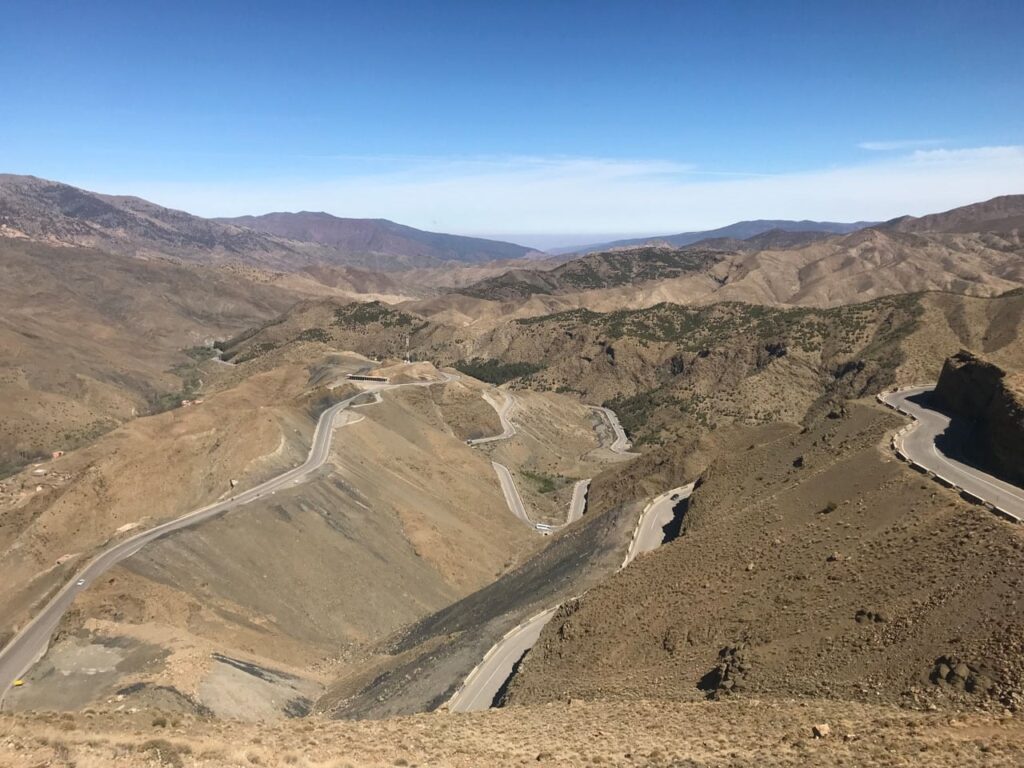 A dramatic winding road with sharp switchbacks through the vast, arid landscape of the Moroccan Atlas Mountains.