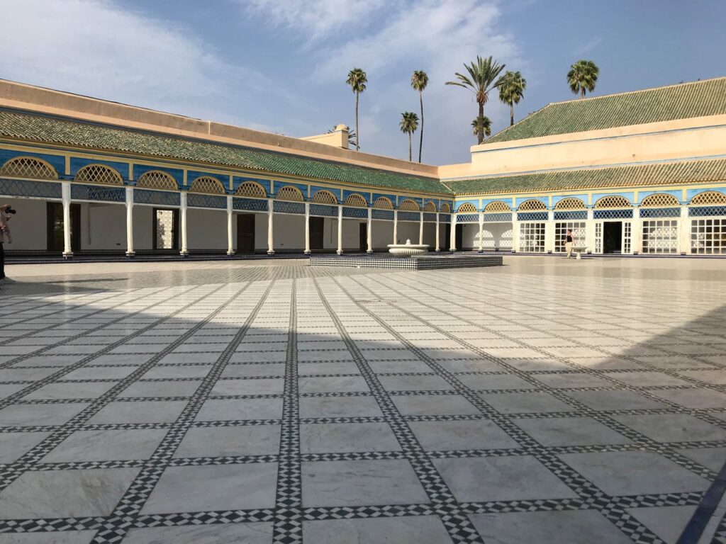 A wide-angle view of a vast, open-air courtyard within the Bahia Palace Courtyard, Marrakesh. The floor is covered in a striking pattern of white and black marble tiles. The courtyard is enclosed by white columns supporting arches decorated with yellow and blue geometric patterns and green-tiled roofs. A small white marble fountain sits in the center, and several tall palm trees rise above the palace walls against a pale blue sky.