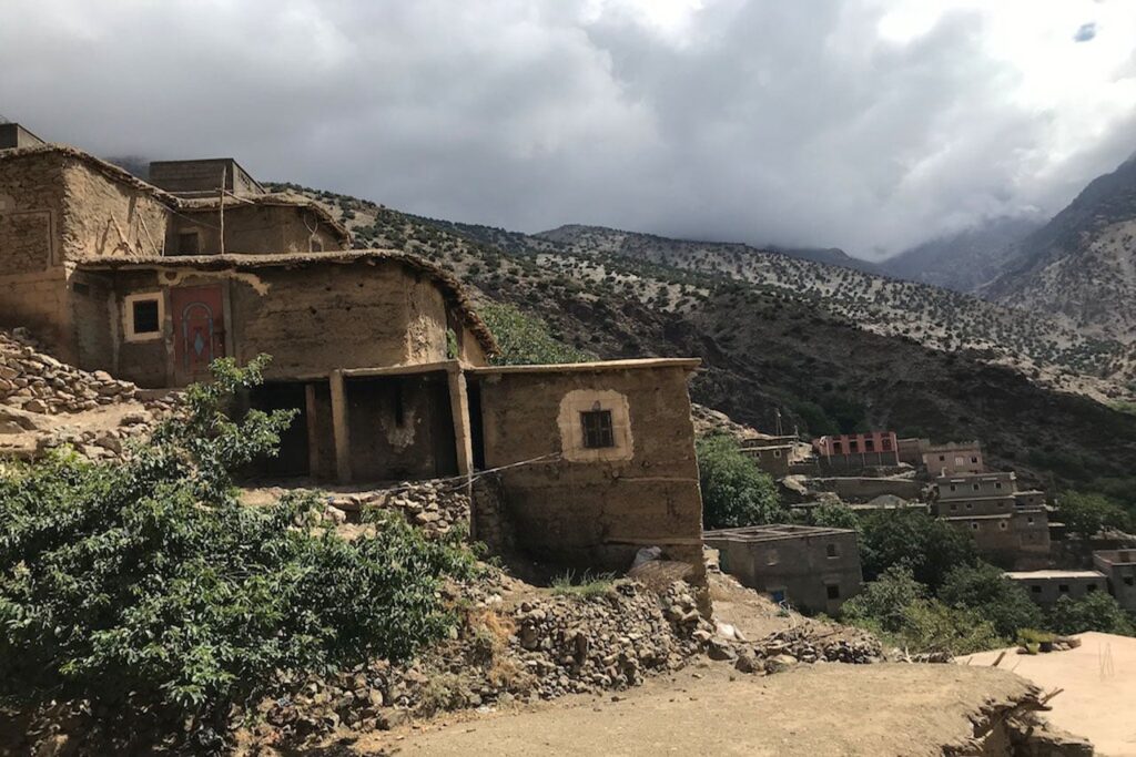 A scenic view of a rustic mountain village in the Atlas Mountains of Morocco. In the foreground, traditional flat-roofed houses made of stone and earth sit on a rocky slope, with a green leafy tree to the left. In the background, rugged, arid mountains rise sharply under a cloudy sky, with more small buildings scattered throughout the valley below.
