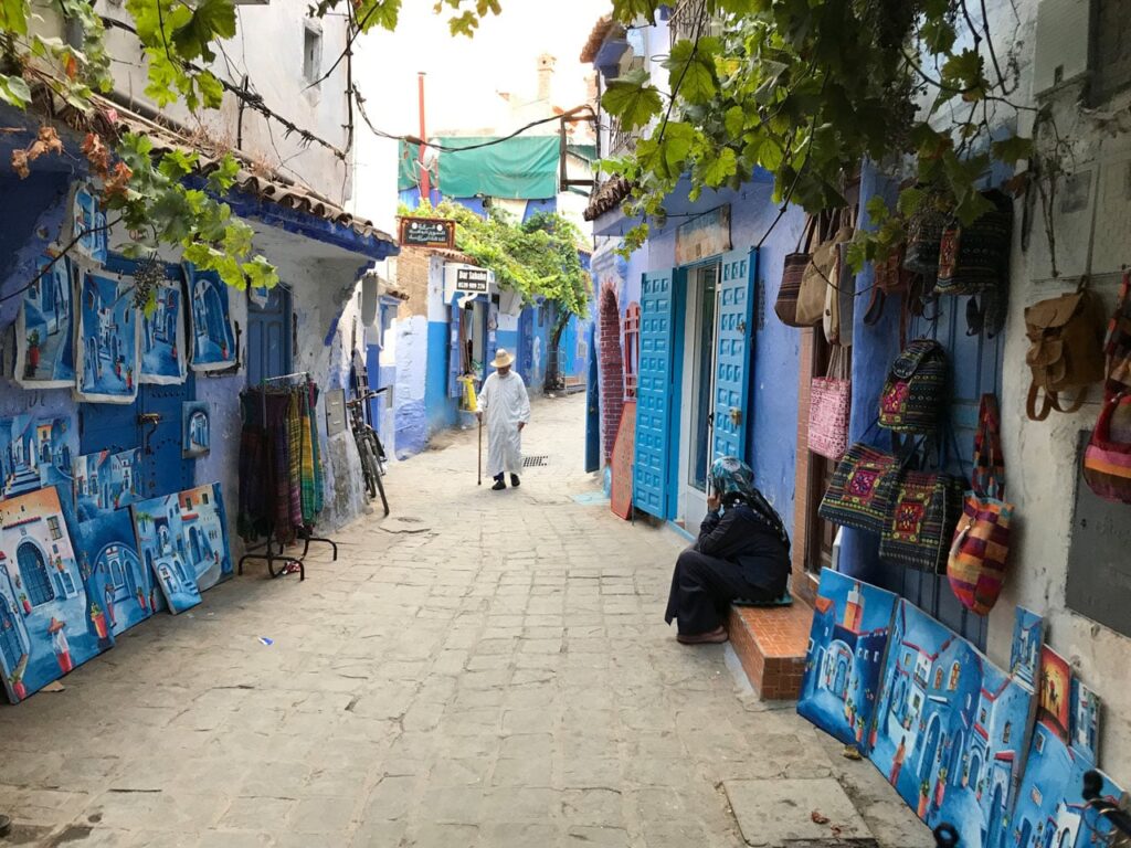 A narrow, cobblestone street in the blue city of Chefchaouen. The walls of the buildings are painted various shades of light and bright blue. An elderly man in a white hooded djellaba walks with a cane in the distance, while a woman in dark clothing sits on a step in the foreground. Blue-themed paintings and colorful woven bags are displayed for sale along the walls, partially shaded by green vine leaves hanging from above.