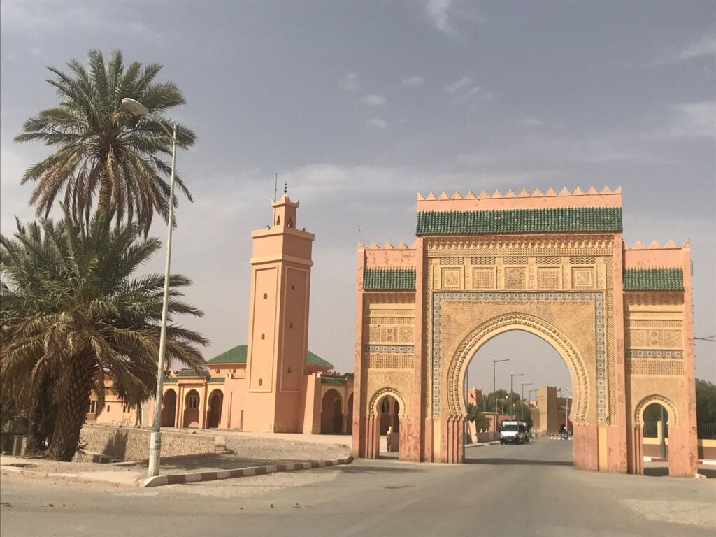 A wide-angle shot of a grand, sandy-pink Moroccan archway featuring intricate geometric carvings and green tiled accents. To the left stands a matching tall, square minaret and a lush palm tree. A road passes through the large central arch, with a white van visible in the distance.