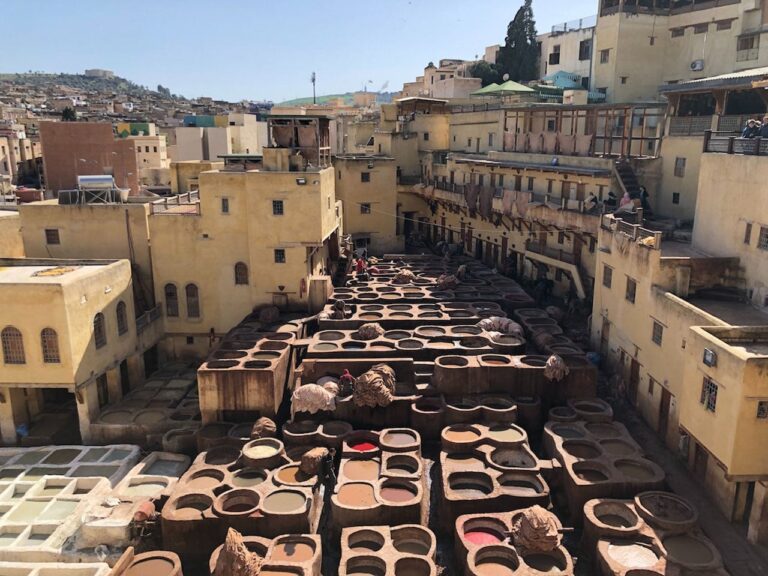 A high-angle view of the Chouara Tannery in Fez, Morocco. The scene is filled with dozens of round stone vats containing various colored dyes and liquids used for processing leather. Workers can be seen moving between the pits. The tannery is surrounded by tall, beige-colored traditional buildings with small windows, and hides are draped over balconies to dry under the bright sun.