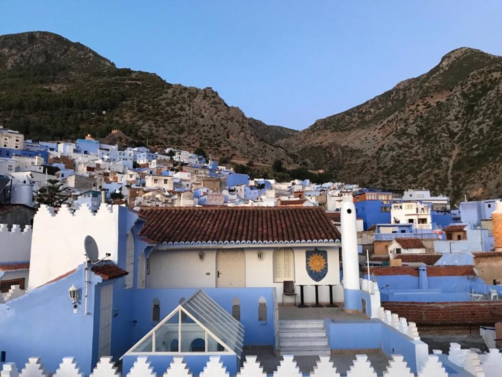 A wide-angle view of the "Blue City" of Chefchaouen nestled at the base of two rugged, steep mountains under a clear sky. In the foreground, white and blue buildings feature terracotta tiled roofs and decorative crenelated walls. A sun mosaic is visible on a white wall. The background shows the densely packed blue-washed houses of the medina climbing up the mountain slope.