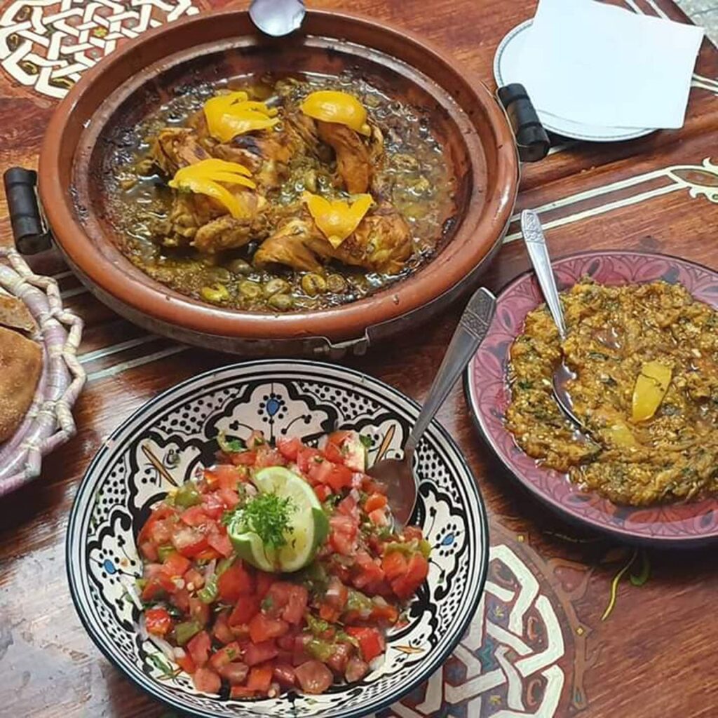 A traditional Moroccan feast featuring chicken tagine with olives, a fresh tomato salad, and eggplant dip served in decorative ceramic bowls.