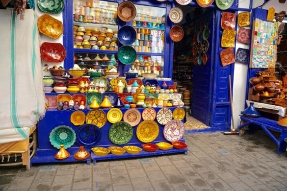 A storefront with a bright blue frame displaying a wide variety of colorful handcrafted Moroccan pottery, including patterned plates, bowls, and conical tagines.