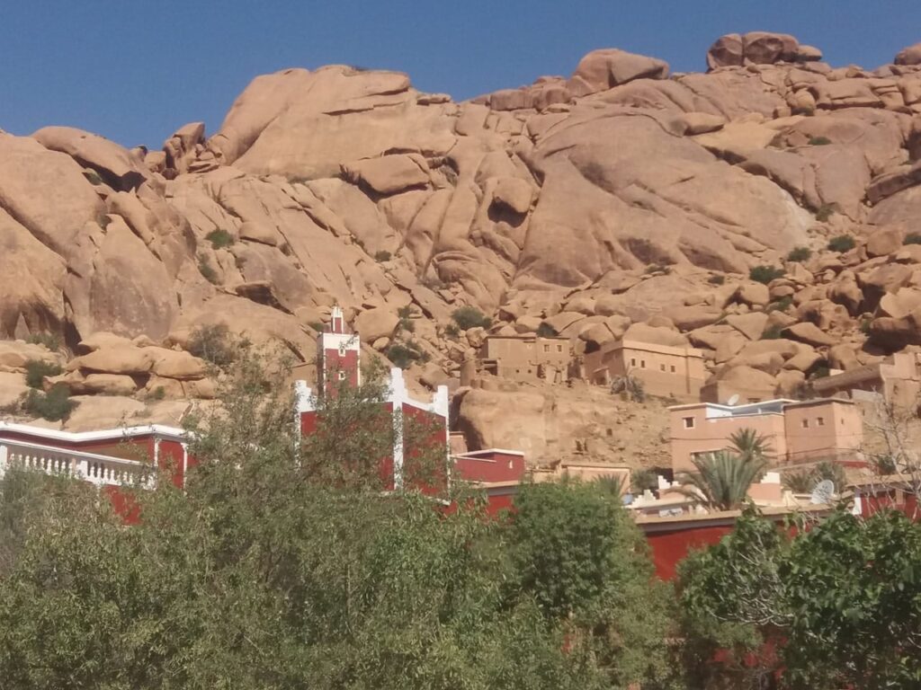 A small village with tan and reddish-brown buildings nestled at the base of a massive, rocky mountain. A tall, slender red minaret with white accents rises above the rooftops, framed by lush green palm trees.