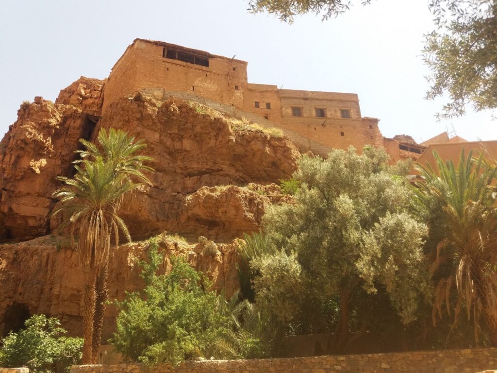 A historic mud-brick building perched on a rocky cliffside, surrounded by green palm and olive trees.