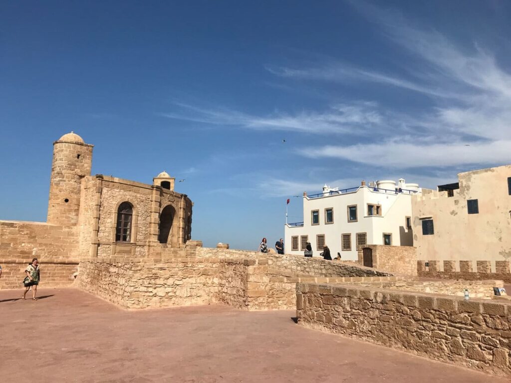 The historic seaside stone ramparts and watchtower of Essaouira, Morocco, set against white buildings and a clear blue sky.