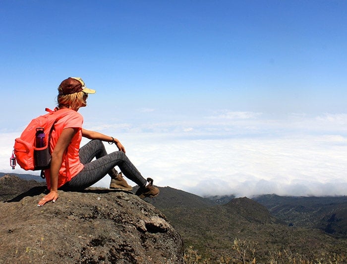 A woman with a bright orange backpack and a hat sits on a rocky cliff edge, looking out over a vast, thick sea of white clouds under a clear blue sky.