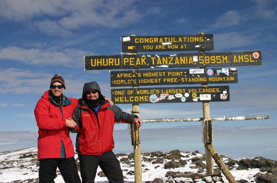 Two male trekkers in red jackets shaking hands and smiling in front of the famous wooden summit sign at Uhuru Peak, Kilimanjaro.