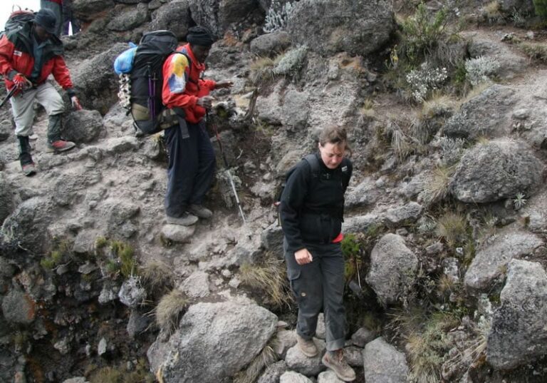 A woman in a black jacket carefully navigates a steep, rocky descent while two guides in red jackets with large backpacks follow behind on the rugged mountain trail.