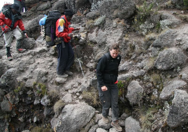 A woman in a black jacket carefully navigates a steep, rocky descent while two guides in red jackets with large backpacks follow behind on the rugged mountain trail.